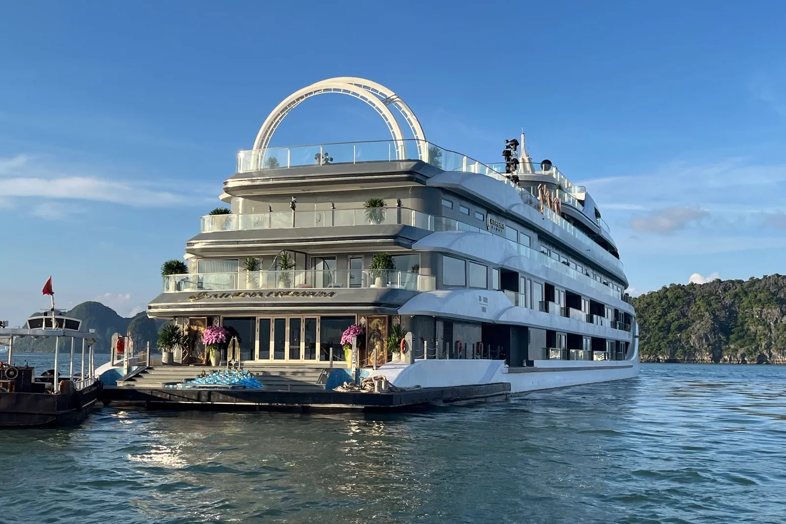 A large multi-deck white cruise ship with an arched structure on its top deck sits on calm water near limestone karst hills, with gold lettering on the hull above the entrance platform and a smaller boat with a red flag at left.