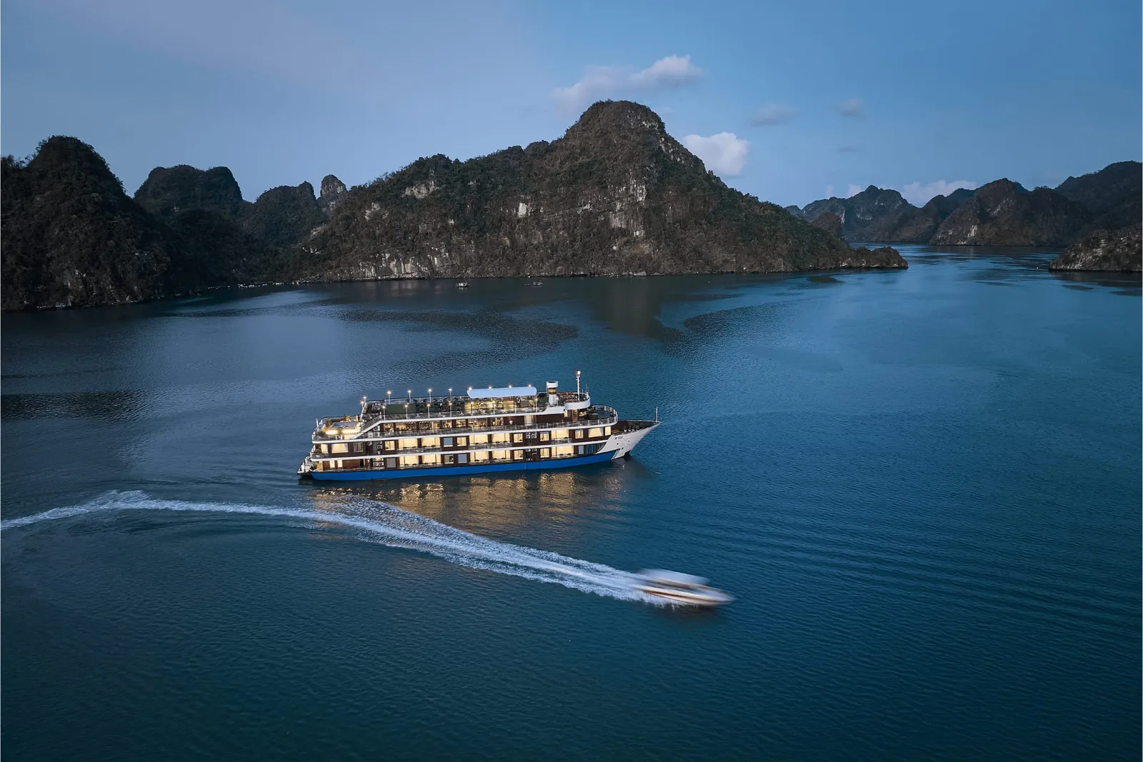 An aerial dusk view of a multi-deck cruise ship with lit windows on calm blue water among forested limestone karst islands, with a speedboat leaving a white wake in the foreground.