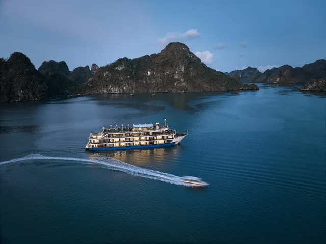 An aerial dusk view of a multi-deck cruise ship with lit windows on calm blue water among forested limestone karst islands, with a speedboat leaving a white wake in the foreground.