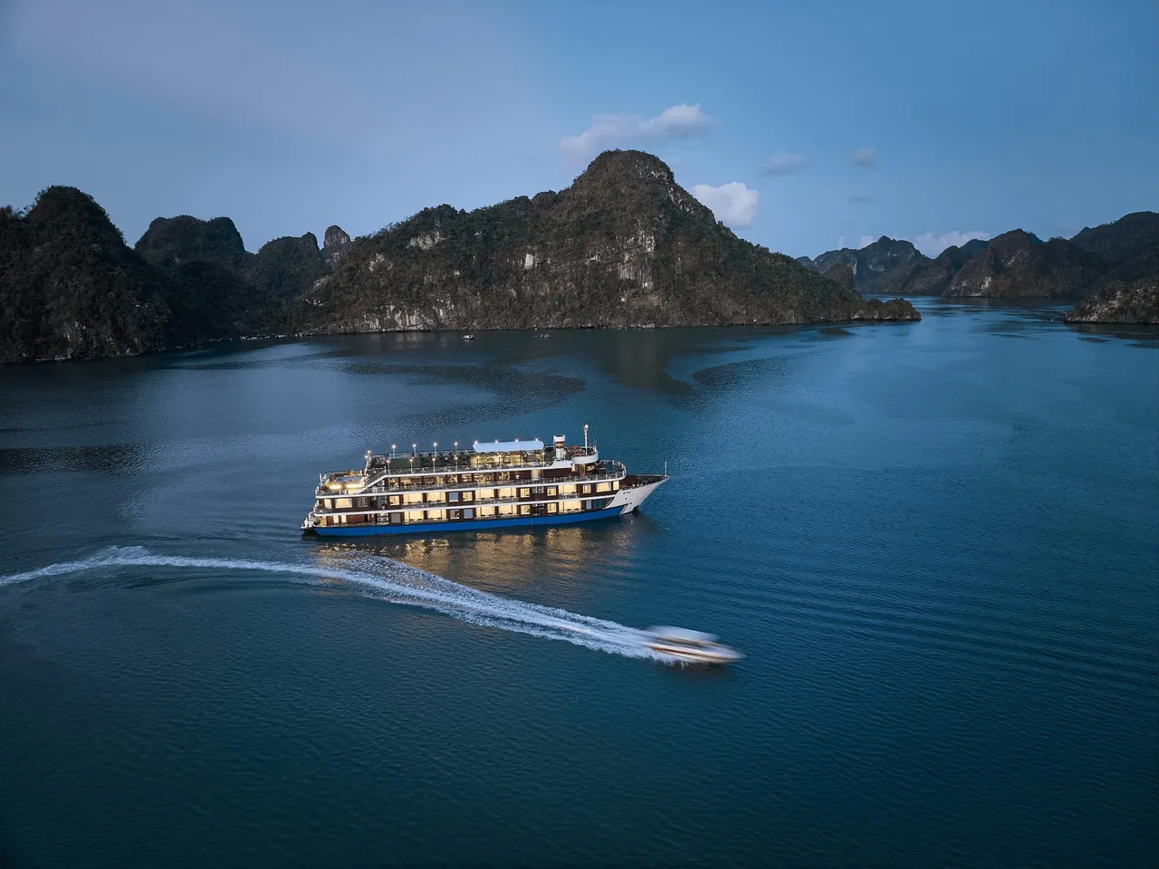 An aerial dusk view of a multi-deck cruise ship with lit windows on calm blue water among forested limestone karst islands, with a speedboat leaving a white wake in the foreground.