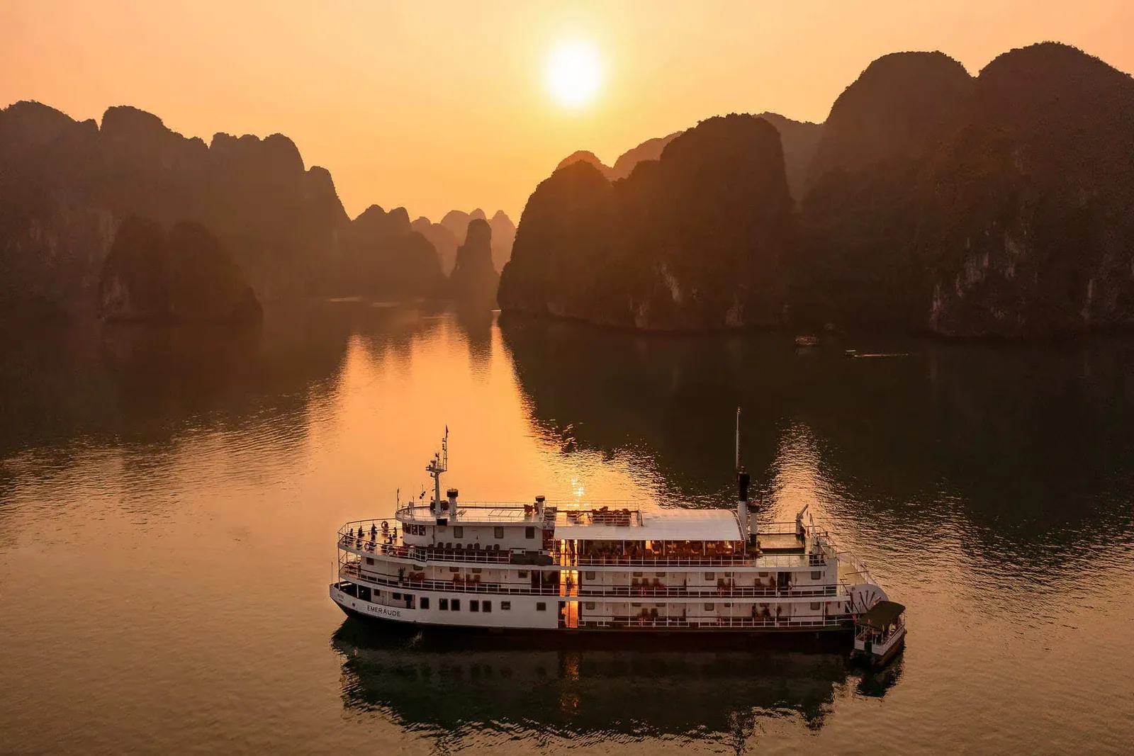 An aerial view at sunset of a white multi-deck paddle-style cruise boat with the name 'Emeraude' on its hull, sailing on calm water among dark limestone karst peaks under an orange sky and low sun.