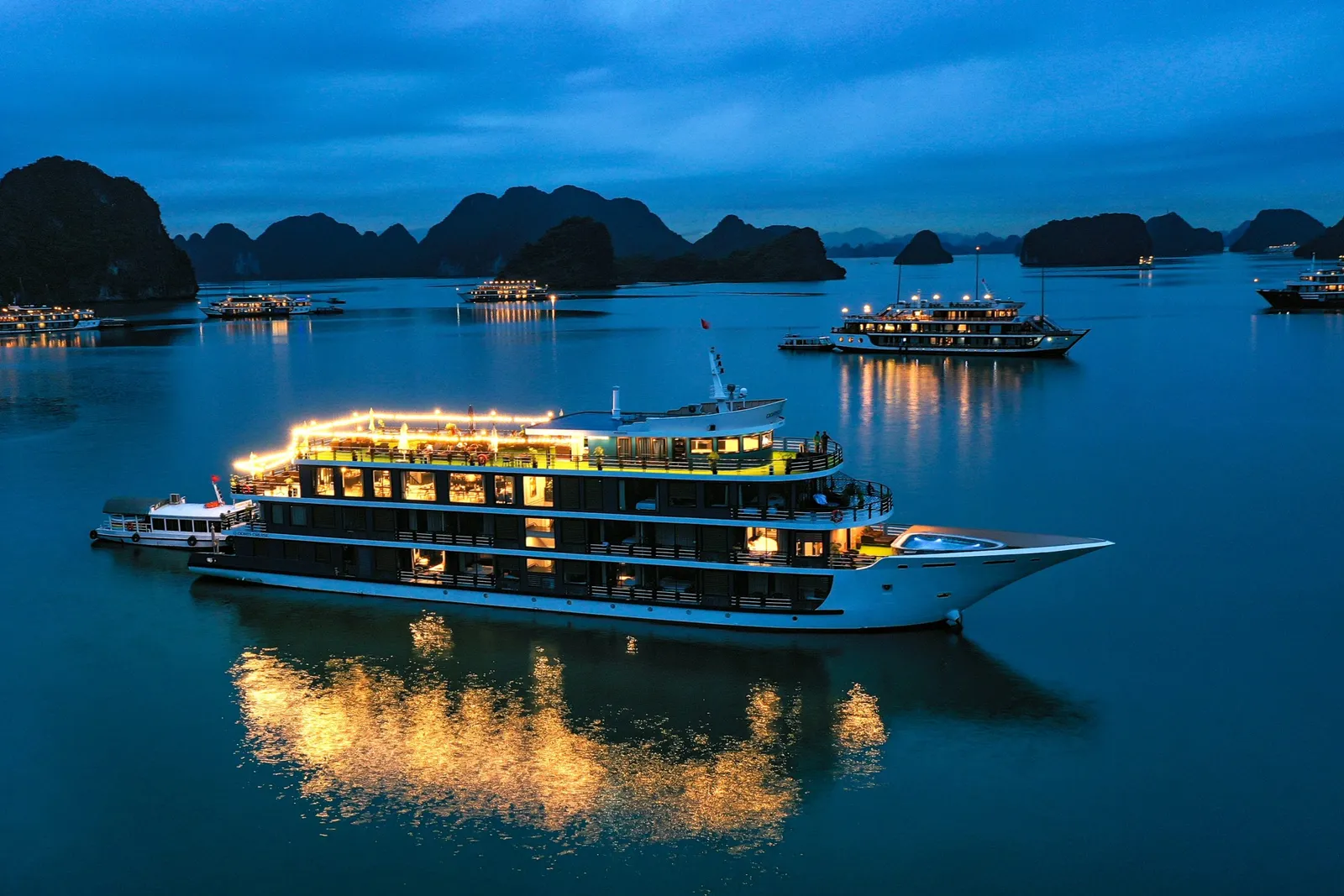 An illuminated multi-deck cruise boat at dusk in a bay surrounded by limestone karst islands with other lit boats in the distance.