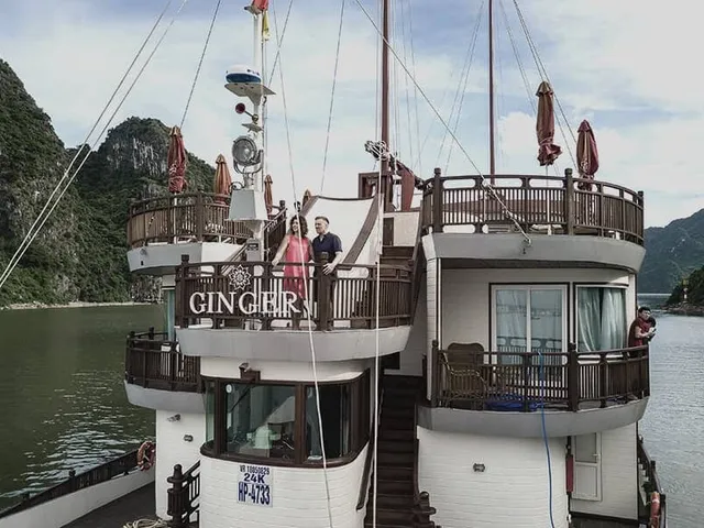 A multi-deck wooden junk boat with 'GINGER' lettering on its balcony railing, a couple standing on an upper deck and another person on a lower deck, with limestone karsts and calm green water around it.