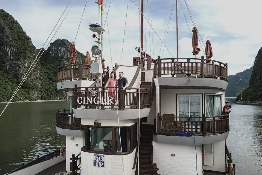 A multi-deck wooden junk boat with 'GINGER' lettering on its balcony railing, a couple standing on an upper deck and another person on a lower deck, with limestone karsts and calm green water around it.