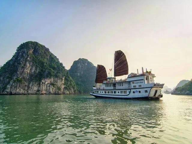 A cream wooden junk with two red sails anchored in calm water in front of green karst islands.