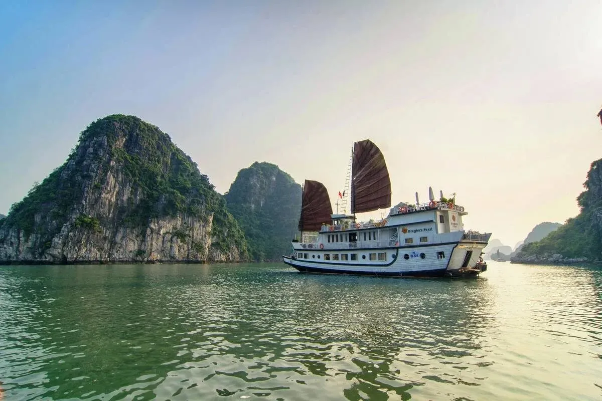 A cream wooden junk with two red sails anchored in calm water in front of green karst islands.