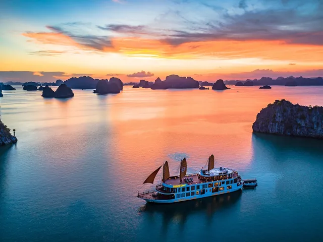 An aerial view at sunset of a blue-hulled cruise ship with orange sails and a lit-up yellow name sign on its hull, floating in calm water among limestone karst islands under an orange and teal sky.