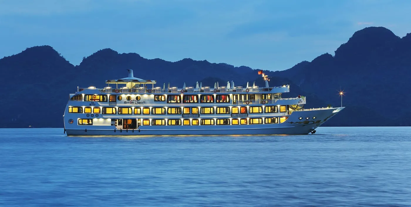 A multi-deck white cruise ship with lit amber windows and the name 'Star Light' on its side sails on calm blue water at dusk in front of dark limestone karst hills, with a Vietnamese flag flying at the top.