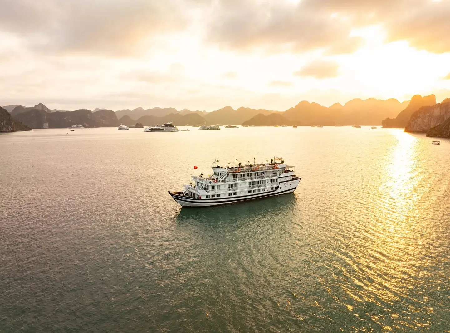 An aerial view at sunset of a white multi-deck cruise boat with a dark striped hull on calm water, with limestone karst islands and other boats in the hazy golden background.