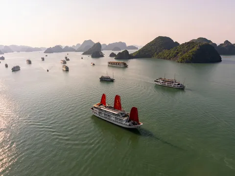 Aerial view of a white cruise boat with three red sails in calm green water, with other cruise boats and forested limestone karst islands in the hazy background.