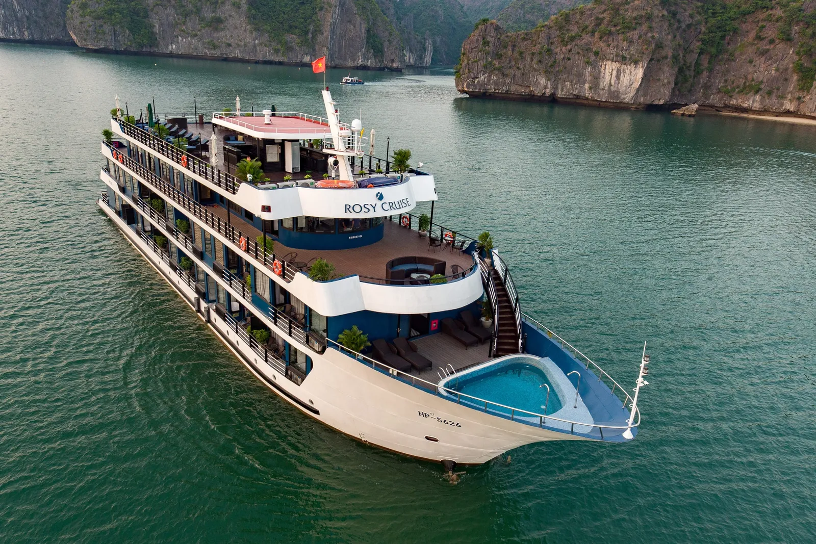 Rosy Cruise broadside on flat green water, four white decks with a plunge pool set into the bow, karst cliffs rising close behind