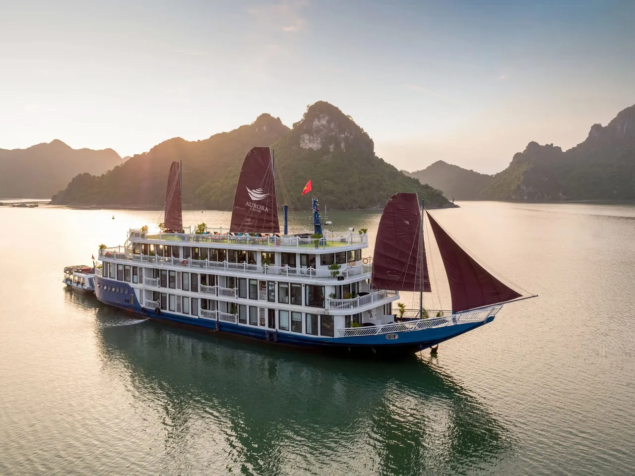 A multi-deck blue-hulled cruise boat with dark red sails, one printed with an 'Aurora' logo, floats on calm water in front of forested limestone karst hills at sunset, with a small tender boat alongside and a Vietnamese flag on top.