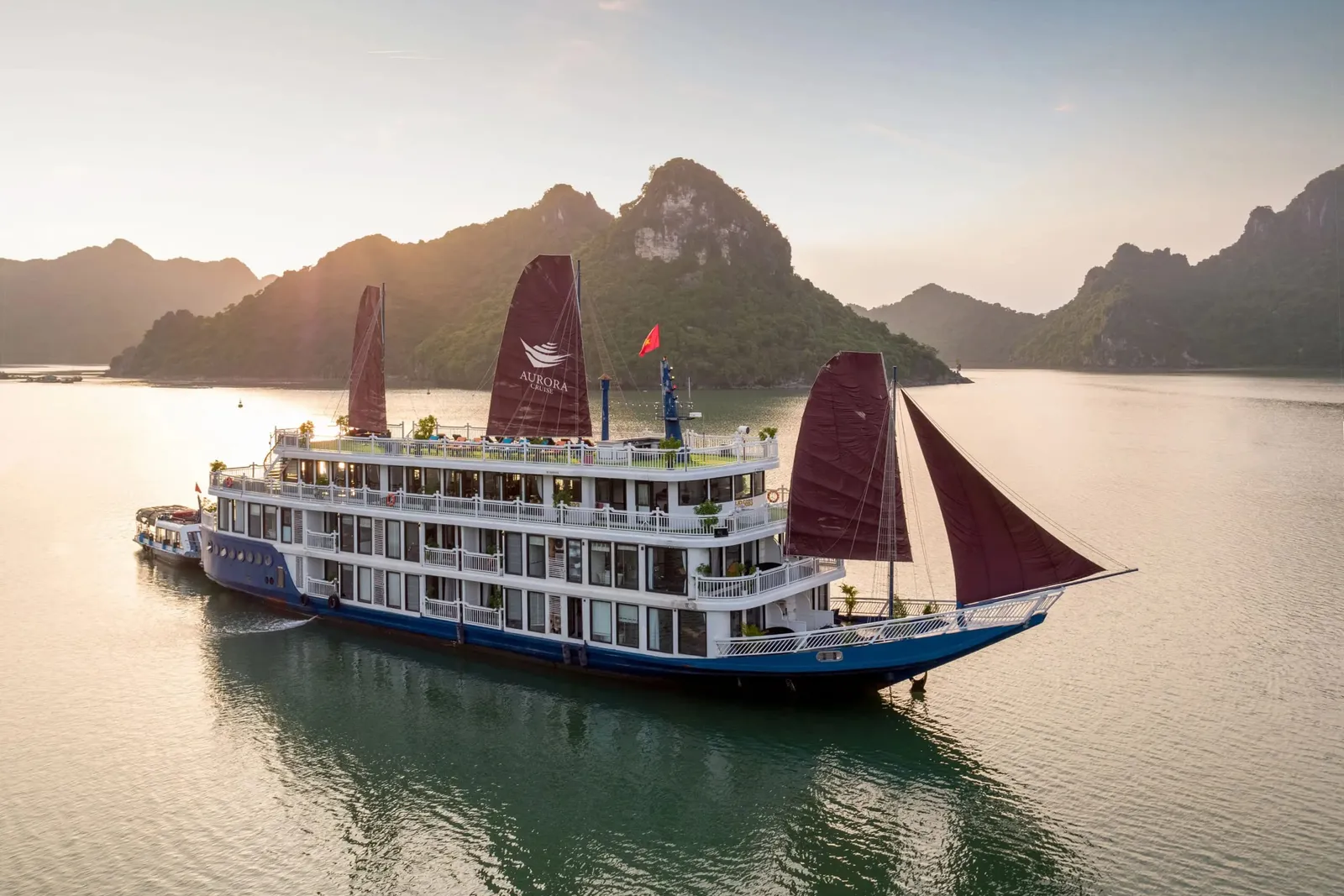 A multi-deck blue-hulled cruise boat with dark red sails, one printed with an 'Aurora' logo, floats on calm water in front of forested limestone karst hills at sunset, with a small tender boat alongside and a Vietnamese flag on top.