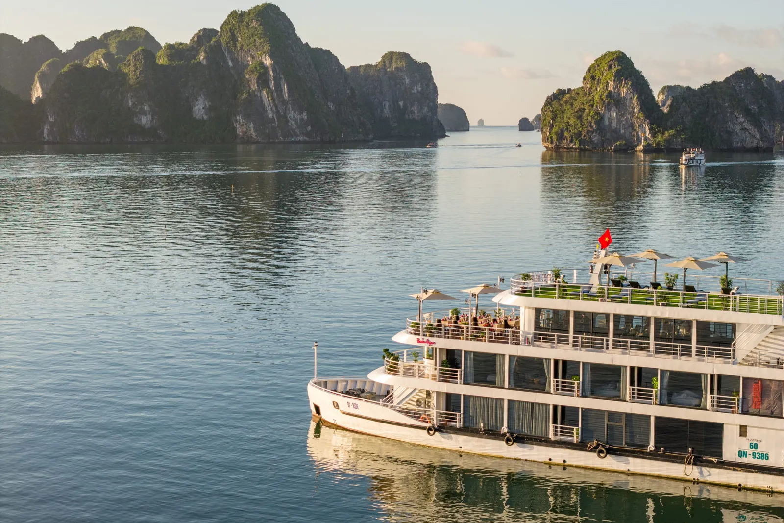 A close aerial view of a white four-deck cruise boat with a dark hull band and hull number QN-9386 on the bow, sailing on calm green water in Ha Long Bay.
