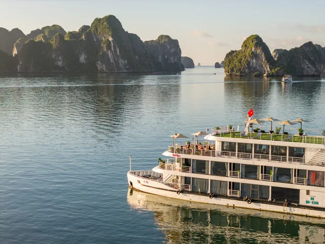 A close aerial view of a white four-deck cruise boat with a dark hull band and hull number QN-9386 on the bow, sailing on calm green water in Ha Long Bay.