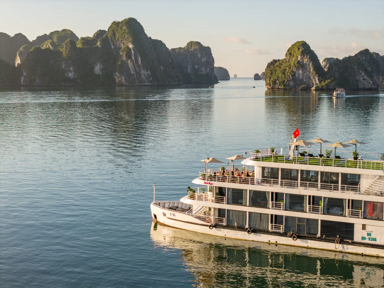 A close aerial view of a white four-deck cruise boat with a dark hull band and hull number QN-9386 on the bow, sailing on calm green water in Ha Long Bay.