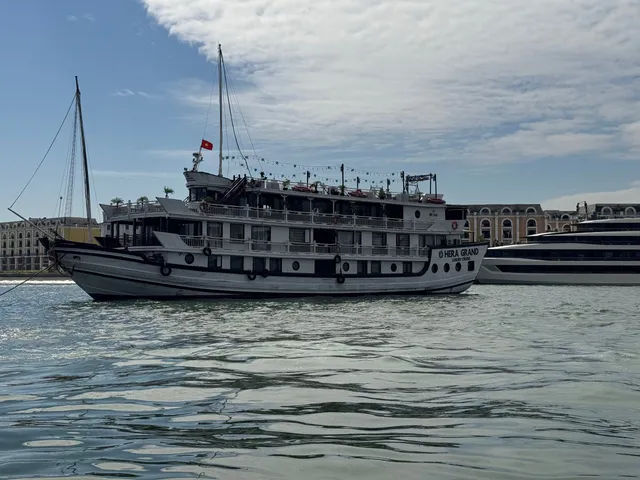 A multi-deck white wooden cruise boat with 'Hera Grand Luxury Cruise' painted on its bow and a Vietnamese flag, photographed from water level, with a larger vessel and arched waterfront buildings behind it.