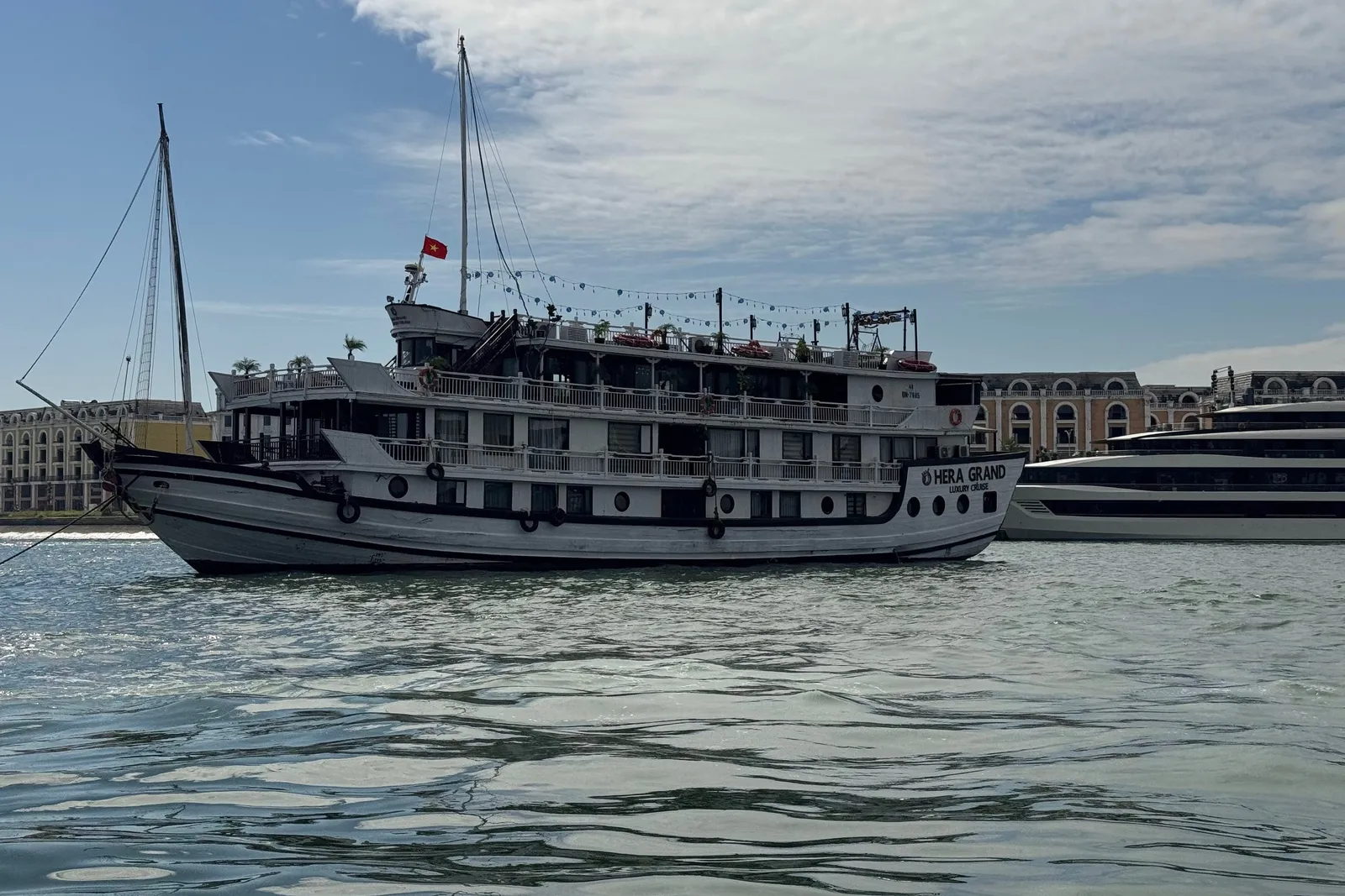 A multi-deck white wooden cruise boat with 'Hera Grand Luxury Cruise' painted on its bow and a Vietnamese flag, photographed from water level, with a larger vessel and arched waterfront buildings behind it.