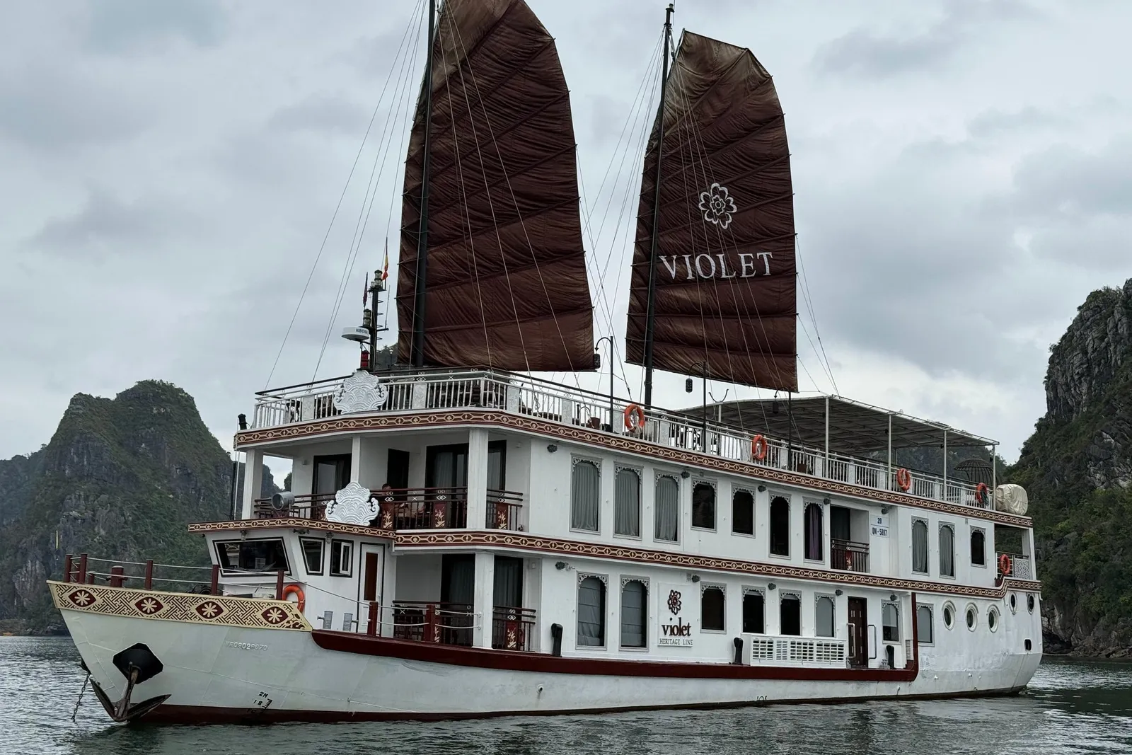 A white multi-deck vessel with two dark brown sails reading VIOLET, anchored among limestone karst islands under an overcast sky.