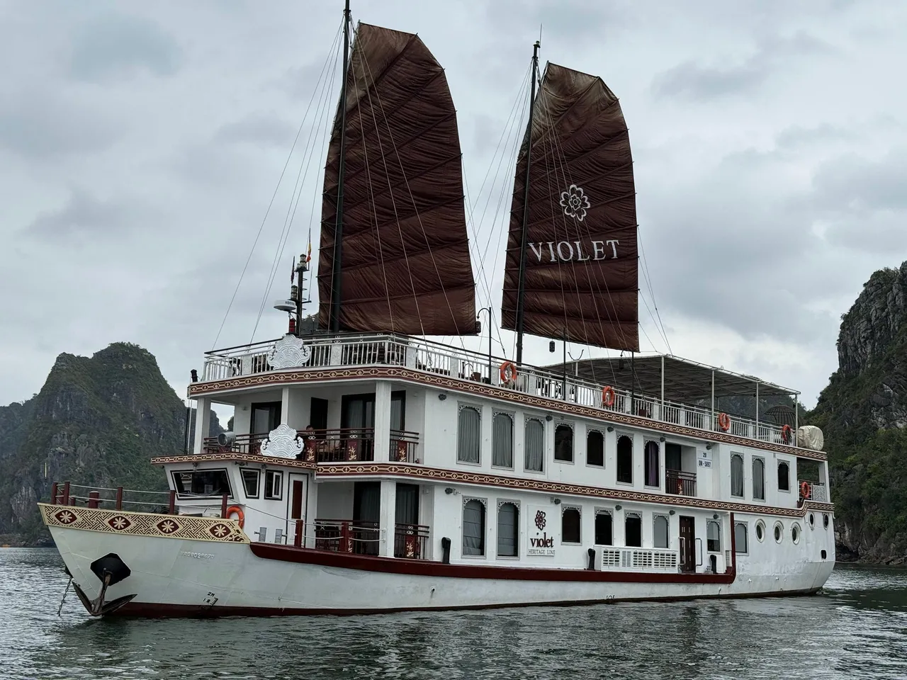 A white multi-deck vessel with two dark brown sails reading VIOLET, anchored among limestone karst islands under an overcast sky.