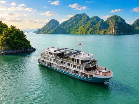 An aerial view of a multi-deck white and blue cruise boat flying a red flag, sailing on turquoise water past forested limestone karst islands under a blue sky with clouds.
