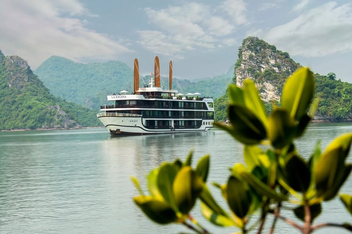 A white multi-deck cruise ship with three orange-brown sails and 'LA REGINA' lettering on its upper hull floats on calm green water among forested limestone karst hills, seen past blurred green leaves in the foreground.