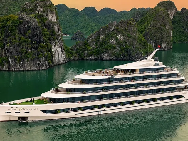 A large white multi-deck cruise ship with a star logo and name on its hull sails on green water past forested limestone karsts under an orange dusk sky, photographed from above and off to the side.