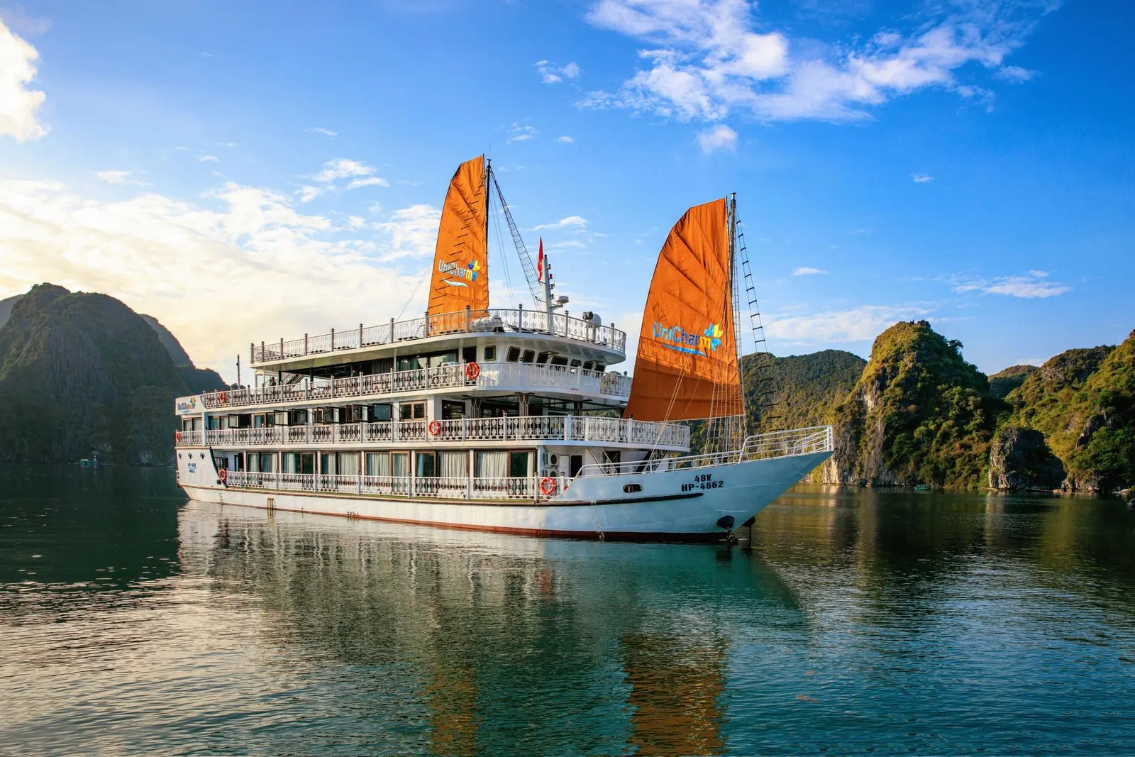 A white multi-deck cruise ship with orange junk-style sails bearing a logo and hull registration text sits on calm green water in front of forested limestone karst islands under a blue sky.
