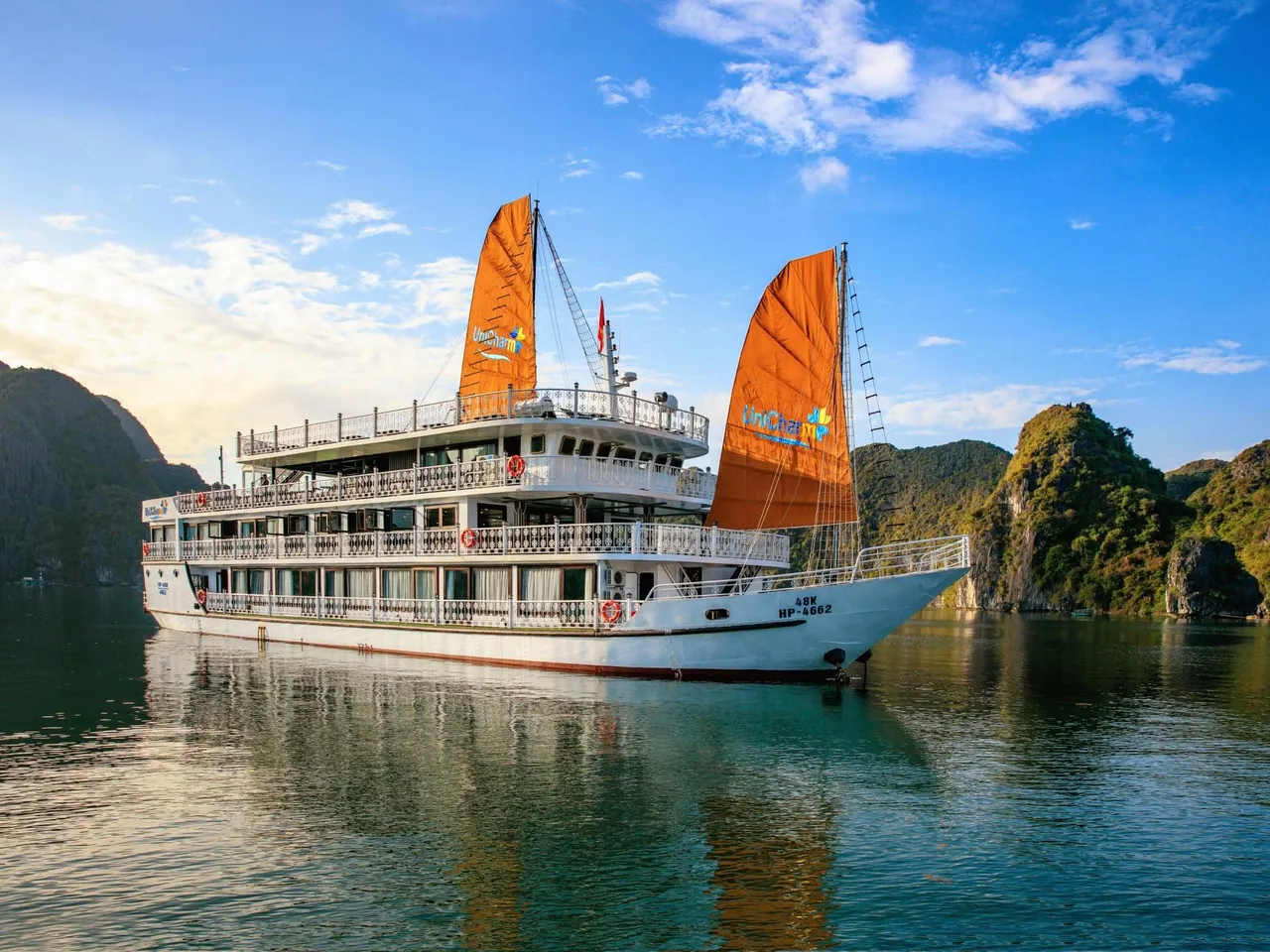 A white multi-deck cruise ship with orange junk-style sails bearing a logo and hull registration text sits on calm green water in front of forested limestone karst islands under a blue sky.