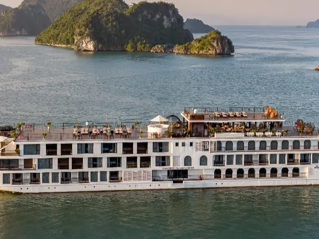 A large white multi-deck cruise ship with a rooftop sun deck sails on calm green water past forested limestone karst islands, with the name painted on the hull near the stern.