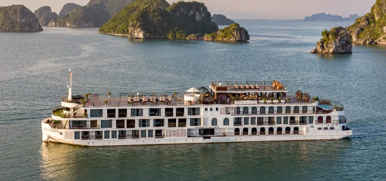 A large white multi-deck cruise ship with a rooftop sun deck sails on calm green water past forested limestone karst islands, with the name painted on the hull near the stern.
