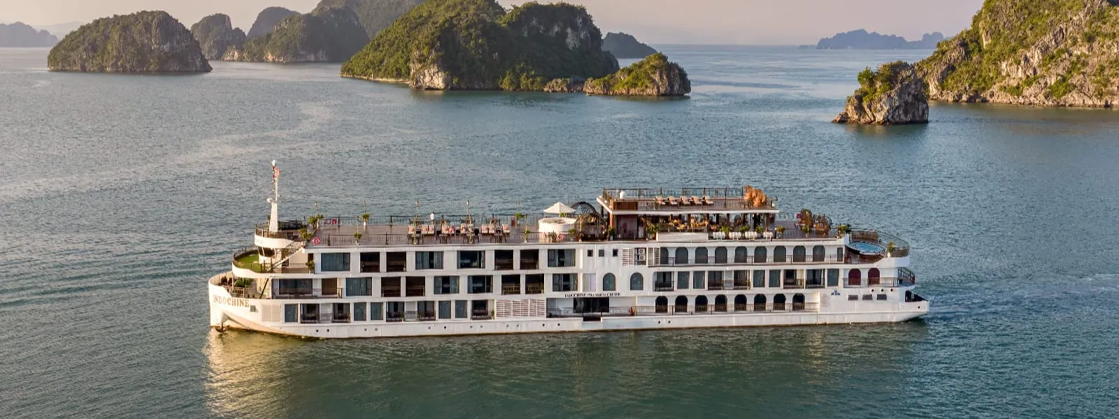 A large white multi-deck cruise ship with a rooftop sun deck sails on calm green water past forested limestone karst islands, with the name painted on the hull near the stern.