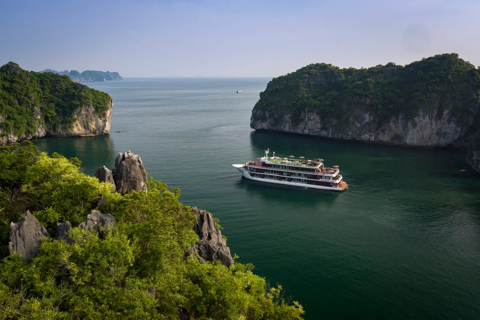A multi-deck white cruise boat sits on calm green water between forested limestone islands, photographed from a high vantage point on a hillside.