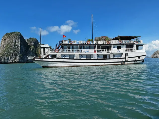 A white cruise boat with multiple decks floats in turquoise water with limestone karst formations in the background under a blue sky.