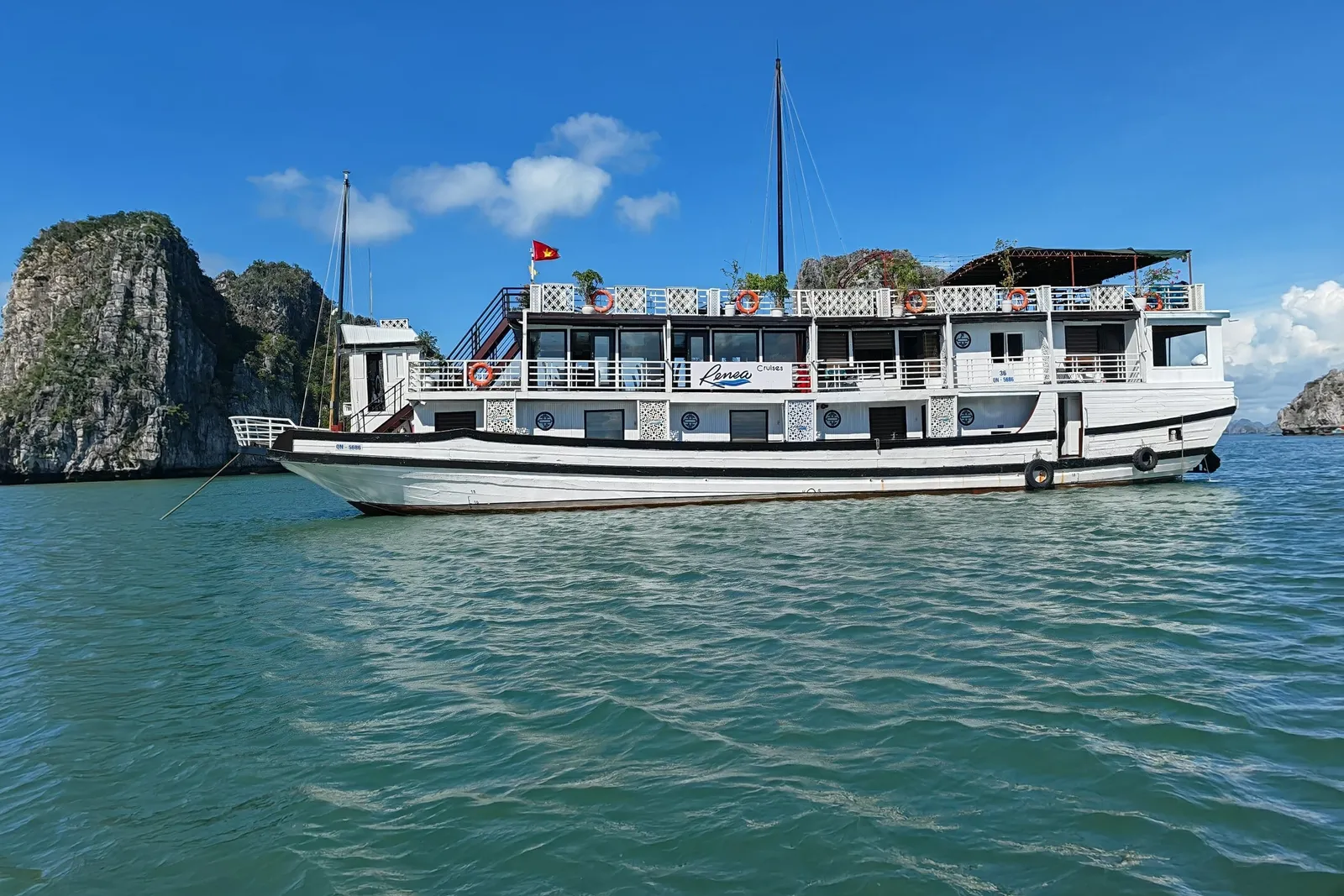 A white cruise boat with multiple decks floats in turquoise water with limestone karst formations in the background under a blue sky.