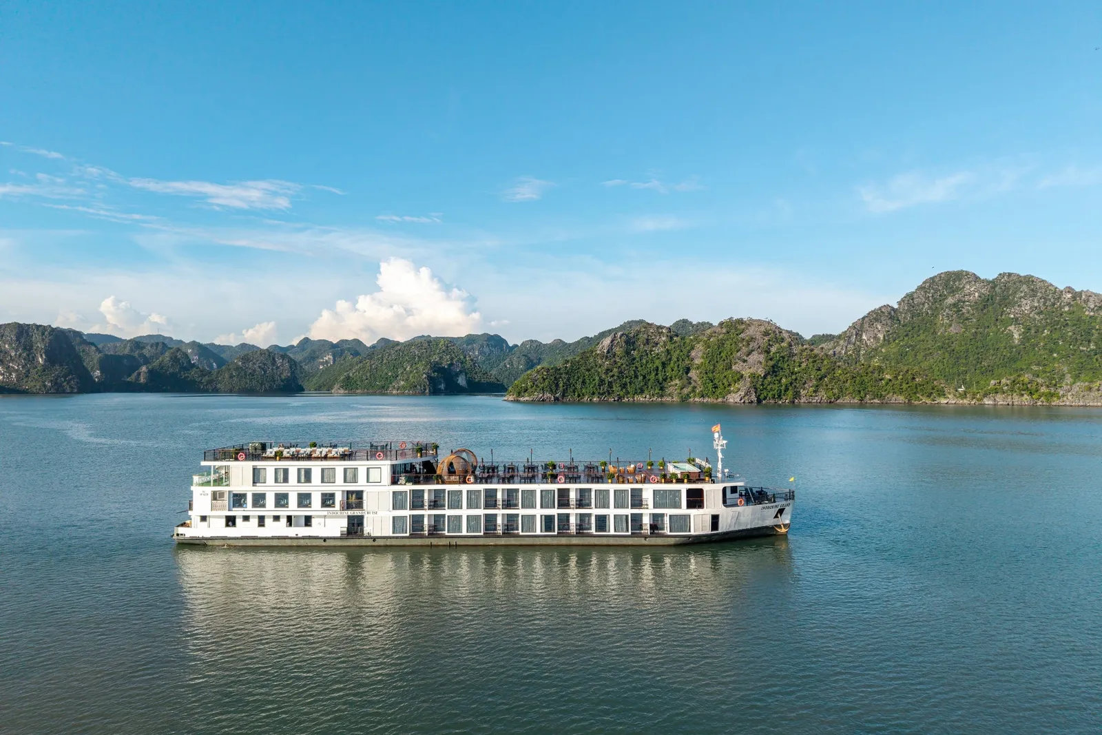 A white multi-deck cruise ship on calm water with limestone karst mountains in the background under blue sky.