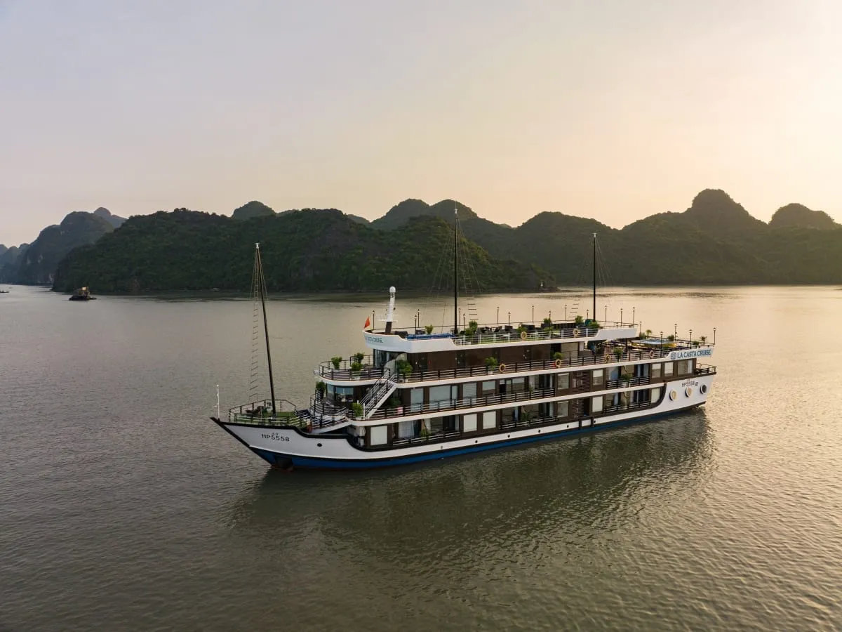 An aerial view of a multi-deck white cruise ship with 'La Casta Cruise' lettering and registration HP 5558 on the hull, anchored on calm water in front of forested limestone karst islands at sunset.