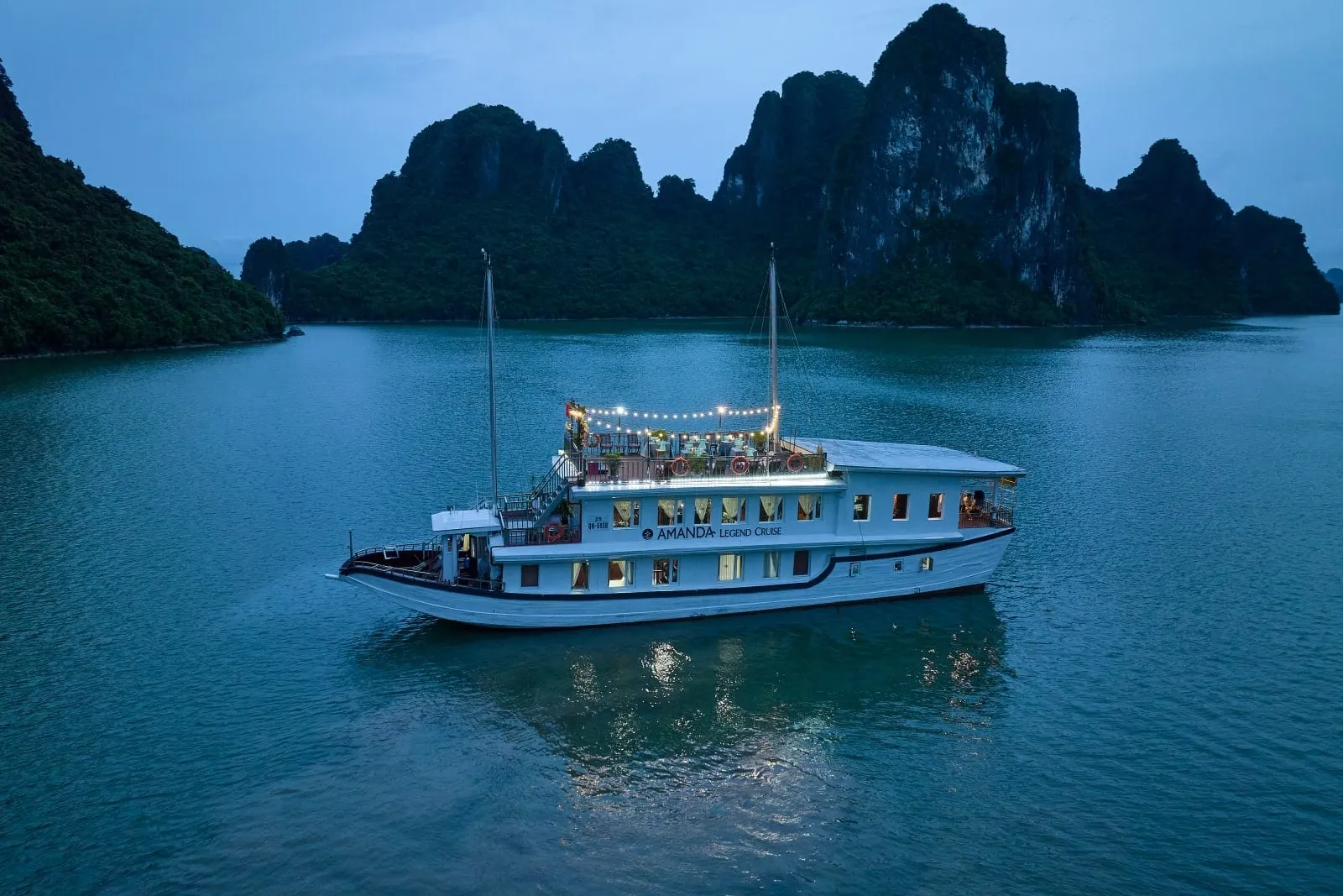 Aerial dusk view of a white two-deck cruise boat with string lights on the top deck, anchored on calm green water in front of forested limestone karsts.