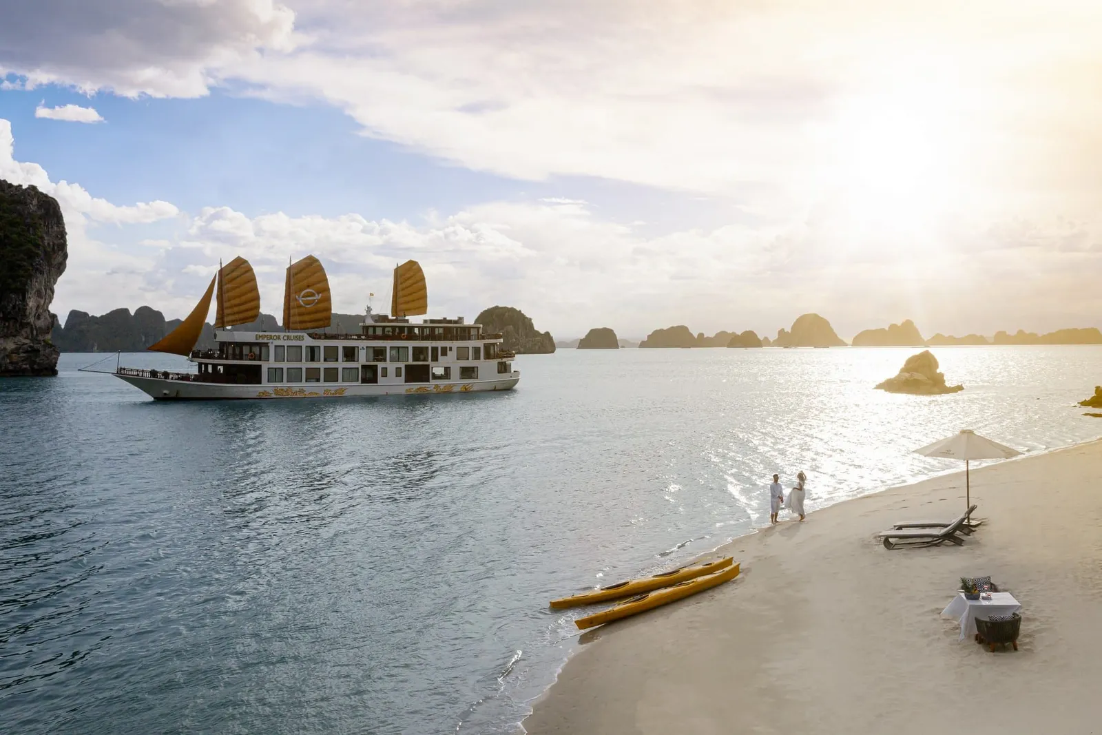 A multi-deck cruise boat with orange junk sails, marked 'Emperor Cruises' on its hull, floats in a bay of limestone karsts at sunset, while two people in white stand on a sandy beach with yellow kayaks, loungers, an umbrella and a small table.