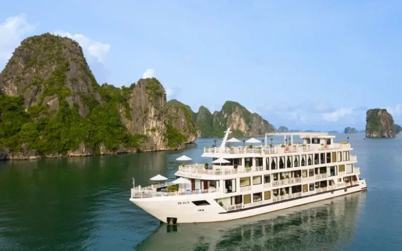 A white multi-deck steel cruise ship with umbrellas on its upper decks sails in calm green water in front of forested limestone karst islands under a blue sky.