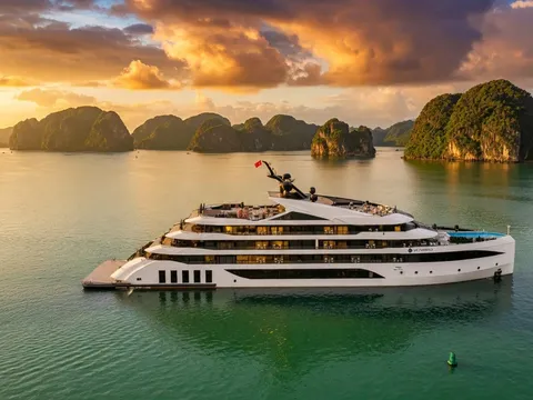 An aerial view of a large white multi-deck cruise ship with small lettering on its hull and a red flag, floating on green water in front of limestone karst islands under a golden sunset sky.