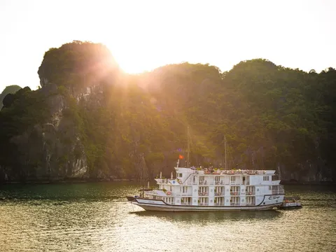 A white multi-deck cruise boat on green water backlit by the sun behind a forested limestone hill.