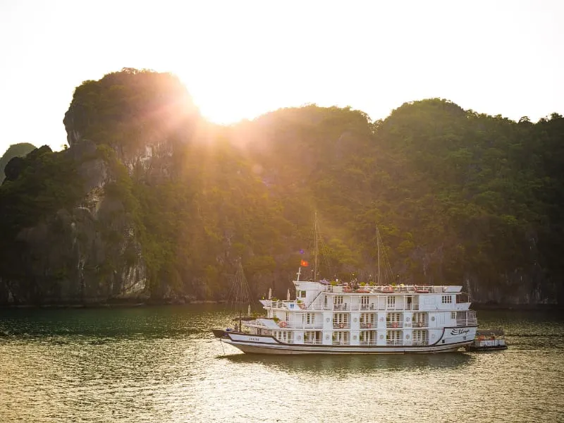A white multi-deck cruise boat on green water backlit by the sun behind a forested limestone hill.