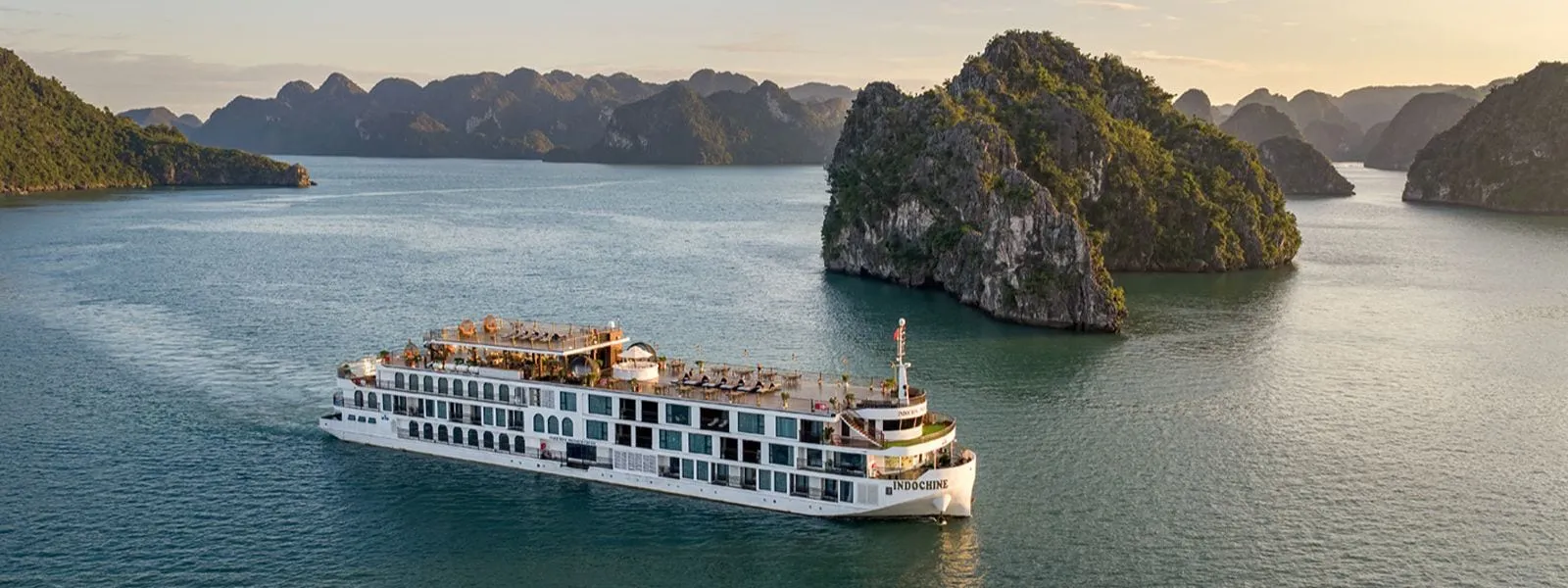 An aerial view of a large white multi-deck cruise ship with the name INDOCHINE on its hull, sailing on calm green water beside forested limestone karst islands at sunset.