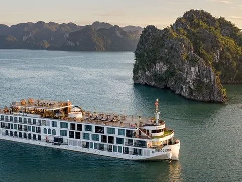 An aerial view of a large white multi-deck cruise ship with the name INDOCHINE on its hull, sailing on calm green water beside forested limestone karst islands at sunset.