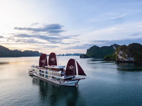 An aerial view of a white multi-deck cruise ship with three dark maroon sails bearing a logo and 'V'Spirit Cruises' text, floating on calm water near green limestone islands at sunset.