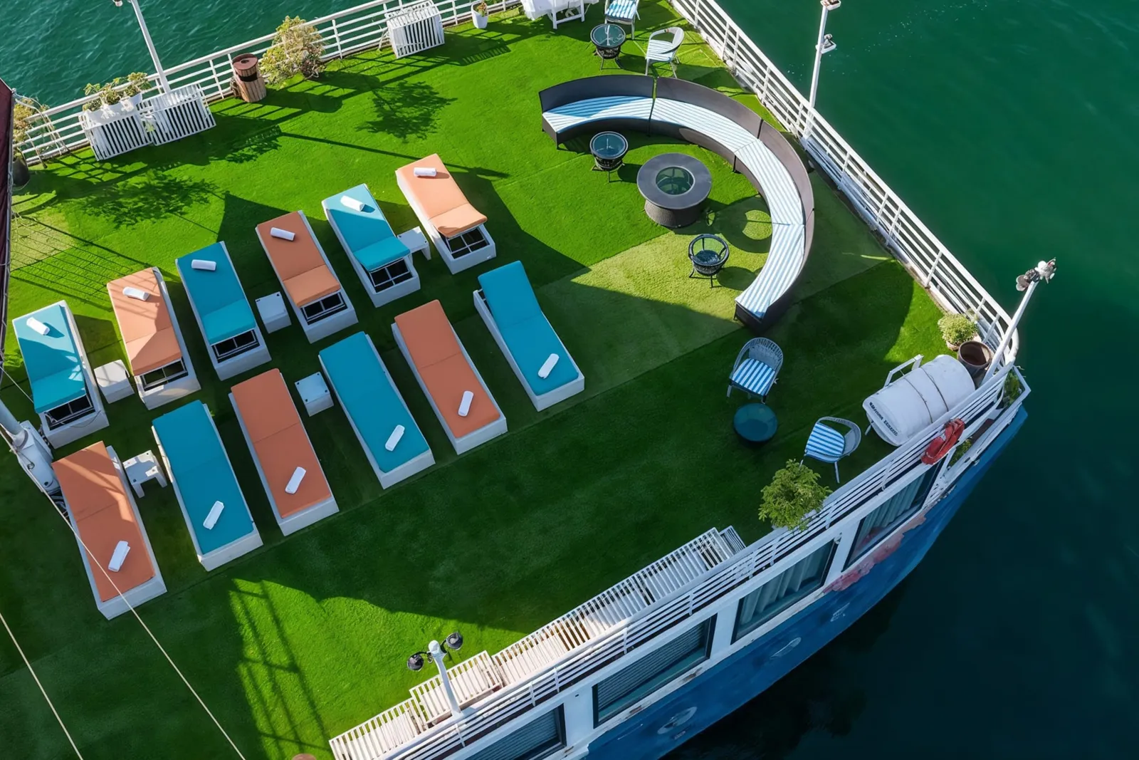 A top-down view of the grass-turf sundeck with orange and teal loungers, with the blue hull edge and a sail visible.