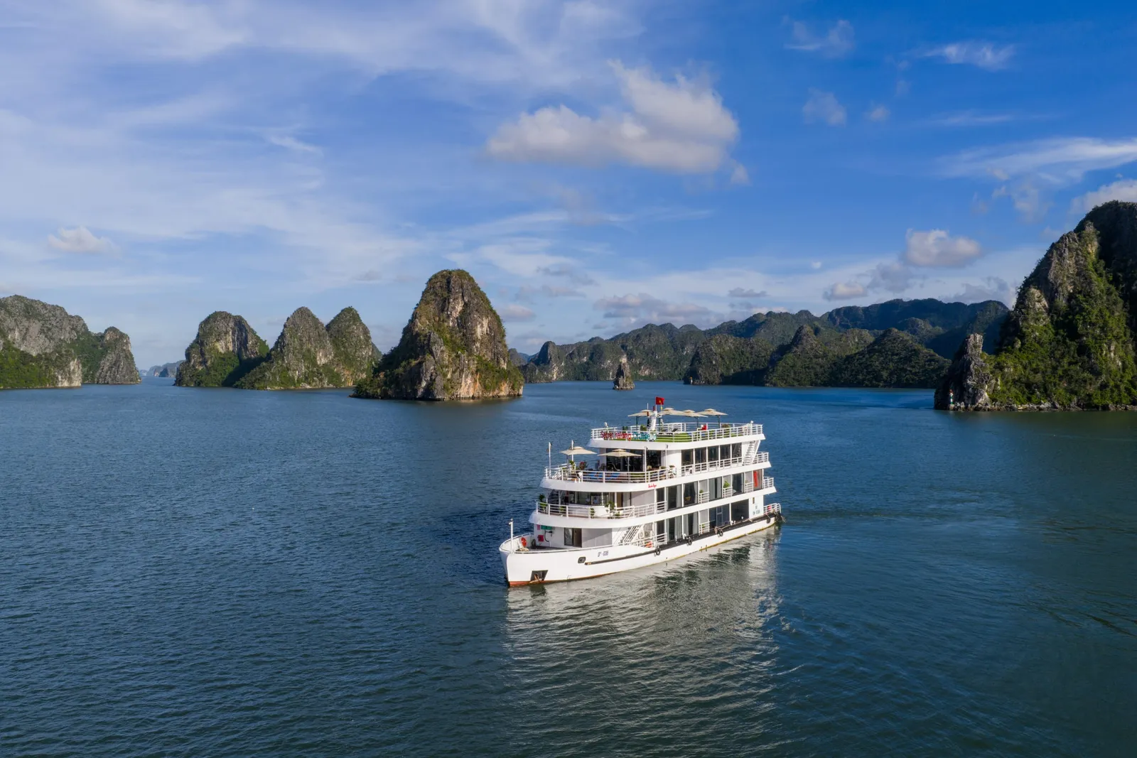 A white multi-deck cruise boat with guests on the upper deck, seen from the water at golden hour with limestone karst islands behind it.