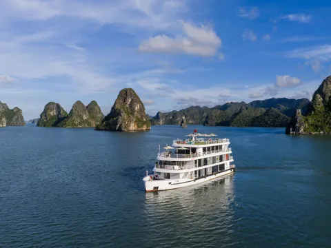 A white multi-deck cruise boat with guests on the upper deck, seen from the water at golden hour with limestone karst islands behind it.