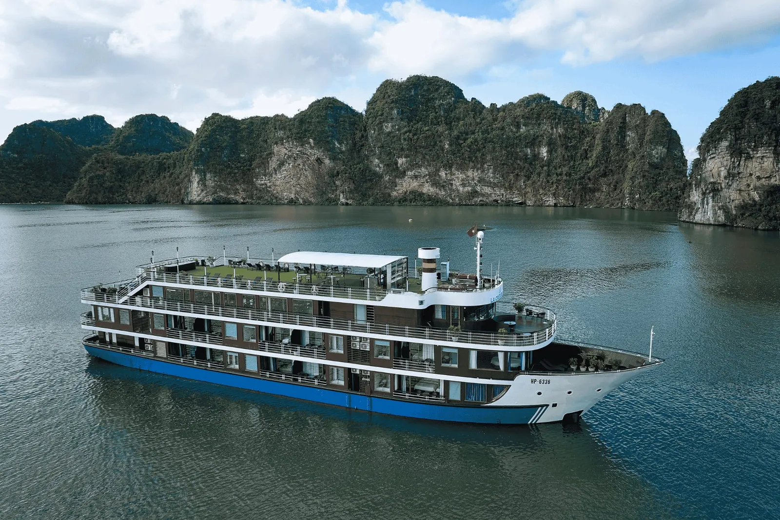 An aerial view of a multi-deck cruise ship with a blue hull, white superstructure and the registration 'HP 6336' on the bow, sailing on calm water in front of forested limestone karst cliffs under a partly cloudy sky.